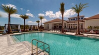 This two-story community amenity center is designed with a resort-inspired aesthetic, blending indoor comfort with open-air leisure. The centerpiece of the outdoor area is a generously sized swimming pool surrounded by a paved sun deck, lounge chairs, and towering palm trees that lend a tropical atmosphere. Draped cabanas and manicured green lawn areas—complete with cornhole boards—offer additional spaces for relaxation and recreation.

The covered indoor-outdoor lounge area features soaring ceilings with large ceiling fans, floor-to-ceiling glass openings that invite abundant natural light and unobstructed views of the pool and grounds. A long bar counter with stone-topped surfaces and woven bar seating anchors one side, while a fully equipped outdoor kitchen with multiple grills serves as a hub for social gatherings. Comfortable seating arrangements with neutral-toned sofas and armchairs are positioned throughout on patterned area rugs.

An adjacent interior lounge showcases soft sage-green accent walls, recessed lighting, a mounted flat-screen display, and casual seating, creating a welcoming retreat. A separate covered game area houses a ping-pong table and foosball table, opening directly toward the pool. The surrounding landscape features well-maintained lawns, paved walkways, and palm-lined grounds that extend across the entire community.