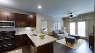 This single-floor residence offers a thoughtfully designed open-concept layout that seamlessly connects the kitchen, living, and sleeping areas. The kitchen is a standout feature, showcasing rich dark-wood cabinetry, a white subway tile backsplash, and a granite-topped island with an undermount sink — providing both style and functionality. The open flow leads into a comfortable living area anchored by warm hardwood-style flooring, a ceiling fan, and a large window that welcomes natural light and offers views of an outdoor balcony area. The living space is complemented by neutral wall tones that lend a calm, welcoming ambiance throughout. Just beyond, the bedroom is visible through an open doorway, featuring soft, neutral décor and ample natural light filtering through window treatments. Crown molding details add a refined architectural touch to the overall interior. The combination of dark cabinetry, granite surfaces, and warm flooring creates a cohesive, modern aesthetic that balances elegance with everyday comfort — making this a practical yet visually appealing living space.