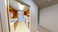This single-floor residential unit offers a practical and functional layout across several well-defined spaces. The galley-style kitchen is lined with warm wood cabinetry on both sides, providing ample storage above and below the counters. It is equipped with a white electric range, a dishwasher, and a stainless steel sink positioned beneath a window that lets in natural light. Light-toned vinyl flooring runs throughout the kitchen, and a ceiling-mounted fluorescent fixture provides bright, even illumination.

The living area is generously proportioned, featuring plush wall-to-wall carpeting and a textured ceiling. A ceiling fan adds comfort, and a baseboard heater runs along the wall for warmth. Large sliding glass doors open directly to a balcony, framing views of surrounding greenery and allowing abundant natural light to fill the room. A wall-mounted air conditioning unit provides additional climate control.

The bedrooms are carpeted and quietly neutral in tone, each with textured ceilings and windows that invite natural light. One bedroom features a baseboard heater and an outlook onto the surrounding landscape.

The bathroom includes a vanity with a dark countertop, white cabinetry below, a large mirror, and bright overhead lighting. The layout is efficient and well-organized, connecting smoothly to the adjacent spaces throughout the unit.