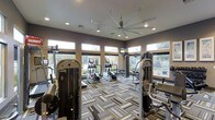 This single-story fitness center is designed to support a full range of workout routines in a bright, welcoming environment. The space is outfitted with an array of strength training machines and cardio equipment, including ellipticals and steppers, strategically arranged to maximize usability and flow. A row of large windows lines the walls, allowing generous natural light to pour in while offering views of the surrounding outdoor landscape, creating an open and energizing atmosphere. Recessed lighting and a large ceiling fan overhead ensure the space remains well-lit and comfortable during all hours of use. The flooring features a distinctive geometric-patterned carpet in neutral tones of gray, beige, and white, adding a modern aesthetic underfoot. Mounted flat-screen televisions provide entertainment options for users during workouts. The walls are finished in warm taupe tones, complementing the contemporary equipment and overall design. The layout is spacious enough to accommodate multiple users simultaneously without feeling crowded, making it a functional and aesthetically refined space for fitness activity.