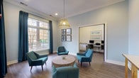This computer room is thoughtfully designed to serve both as a functional workspace and a comfortable lounge area. The space is finished with soft blue-gray walls, crown molding, and wood-look tile flooring throughout, creating a cohesive and polished environment.

The main lounge area is anchored by a round wood coffee table surrounded by mid-century modern teal accent chairs, offering a relaxed seating arrangement beneath a striking gold wire pendant light. Large windows dressed with navy curtain panels flood the space with natural light and provide views of the outdoor surroundings, enhancing the open, airy feel of the room.

A dedicated workstation room is accessible through a wide doorway and features a built-in granite-topped desk spanning the full width of the wall, paired with white cabinetry below for storage. Two monitors, a shared printer, and comfortable upholstered office chairs make this area fully equipped for productivity. Framed landscape artwork adorns the walls above the workstation, adding a calming visual element.

A connecting corridor, lined with a slim console table and wall-mounted TV bracket, links the spaces and leads toward additional areas. The overall design balances functionality with a refined, welcoming ambiance suitable for work or casual use.