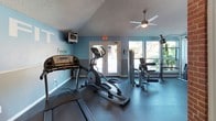 This fitness center offers a thoughtfully designed space built to support an active lifestyle. The facility is divided into distinct functional areas, each serving a specific purpose for residents or members. The main workout area features durable rubber flooring and is outfitted with cardio equipment including treadmills and an elliptical trainer, as well as strength training machines and free weights. Vaulted ceilings with a ceiling fan add an open, airy feel to the exercise space, while large windows and glass-paneled doors allow ample natural light and views of the surrounding greenery. Motivational words such as "Strength," "Focus," "Goal," and "Fit" are displayed on the walls, reinforcing the energetic atmosphere. Exposed brick walls lend a warm, industrial character to the interior, creating a visually distinctive environment. A separate enclosed workout room, accessible through white-framed glass doors set within brick columns, provides additional equipment for targeted training. Adjacent to the fitness area is a business center featuring hardwood-style flooring, vaulted ceilings, workstation desks with computers, and a printer station — ideal for residents needing a quiet, productive workspace. Together, these spaces create a well-rounded amenity facility that balances physical fitness with practical convenience.