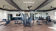 Located at 214 Kingsmeadow Cir in Belville, NC, this fitness center offers a comprehensive workout environment spread across a single floor. The space is divided into two distinct exercise rooms connected by hallways, together providing a generous and well-organized facility for a full range of training activities.

The main exercise room is the heart of the facility, featuring an impressive array of strength training machines, cable systems, free weights, and benches. A wall of mirrors amplifies the sense of space and allows for proper form monitoring, while built-in dark cabinetry provides convenient storage. The room is crowned with a distinctive textured coffered ceiling accented by bold dark trim, ceiling fans, and recessed lighting, lending a modern, upscale atmosphere. A wall-mounted flat-screen TV adds an entertainment element to workouts.

The second exercise room is dedicated to cardio and cycling, housing treadmills, ellipticals, rowing machines, and Peloton bikes. Large black-framed windows flood this area with abundant natural light and offer views of the surrounding outdoor amenity spaces, including a glimpse of a pool area. A wall-mounted TV and stylish patterned wallcovering give this room a boutique fitness studio feel.

Throughout both rooms, durable flooring, ample natural light, and thoughtful design details create a motivating and polished environment well-suited for serious fitness pursuits.