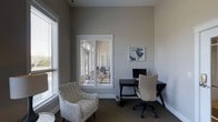 This single-floor space offers a thoughtfully designed environment that balances productivity and comfort. The office area is anchored by a sleek dark desk paired with a cushioned ergonomic chair, complemented by a monitor setup ready for work. A stylish patterned accent chair provides an inviting spot for reading or casual meetings, adding a touch of warmth to the room's professional atmosphere. The walls are finished in a soft, neutral gray tone that creates a calm and focused ambiance throughout the space. White-trimmed windows dressed with horizontal blinds line the room, allowing generous amounts of natural light to filter in while maintaining privacy and light control. The windows also offer views to an adjacent outdoor area, further enhancing the open, airy feel of the interior. Subtle wall art adds a decorative accent without distracting from the clean, understated aesthetic. The carpeted flooring contributes to a quiet, comfortable setting, while track lighting overhead ensures the space remains well-lit during any hour. Overall, this space presents a refined, functional environment well-suited for focused work or professional use.