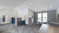 Located at 5600 Briar Oaks Ln in Raleigh, NC, this single-floor office space offers a thoughtfully designed work environment that balances open collaboration with focused productivity. The interior showcases a contemporary aesthetic with vaulted ceilings, recessed lighting, and a neutral color palette of crisp whites and cool grays that creates a clean, professional atmosphere throughout.

The workspace is outfitted with modern open-plan workstations featuring sleek white desks and ergonomic seating, ideal for team-based work. Freestanding acoustic privacy panels are strategically positioned throughout the floor to provide sound separation and visual privacy without sacrificing the open feel of the space. A casual seating area with vibrant blue upholstered sofas adds a welcoming lounge element, perfect for informal meetings or collaborative discussions.

Large multi-pane windows flood the workspace with generous natural light while offering pleasant views of the surrounding greenery, contributing to a bright and inviting ambiance. Textured gray carpet tiles cover the floors, adding both visual interest and acoustic dampening. A coat rack and thoughtfully placed artwork provide finishing touches that make the space feel functional yet personable. Overall, this office is well-suited for dynamic teams seeking a modern, adaptable work environment.