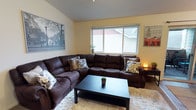This single-floor space offers a warm and welcoming atmosphere centered around a thoughtfully arranged living area. The room features vaulted ceilings that add a sense of height and openness, complemented by soft yellow walls that create a bright, cheerful ambiance. A large window allows generous natural light to fill the space while offering a view of the surrounding exterior.

The living area is anchored by a spacious dark brown leather sectional sofa, generously sized to accommodate multiple guests and adorned with patterned accent pillows. A dark rectangular coffee table sits atop a plush, cream-colored shag area rug, adding texture and comfort underfoot. The combination of dark furniture against the lighter walls and rug creates a balanced, cozy contrast.

Decorative wall art — including cityscapes and urban photography prints — adds character and a cosmopolitan touch to the room. A small side table with a warm amber lamp provides a soft accent light in the corner. A petite potted plant on the windowsill and another on the coffee table bring subtle natural elements indoors. Overall, the space is clean, functional, and designed for comfortable everyday living.