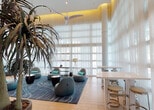 Located at 2263 S Douglas Rd in Miami, FL, this sophisticated lobby lounge offers an impressive and welcoming interior defined by its soaring double-height ceilings and expansive floor-to-ceiling sheer white curtains that diffuse abundant natural light throughout the space. The open-plan layout is anchored by light wood flooring and a large, custom area rug featuring an abstract wave-like pattern in deep teal and neutral tones, evoking Miami's coastal character.

The seating area is thoughtfully arranged with contemporary pod-style lounge chairs in soft gray upholstery, each accented with teal throw pillows that reinforce the space's coastal color palette. Sculptural side tables in both dark matte and geometric wood-inlay finishes are interspersed throughout, complementing the organic forms of the furnishings. Tall tripod floor lamps provide warm, ambient lighting alongside recessed ceiling fixtures and a modern ceiling fan.

A counter-height communal table with sleek bar stools offers an alternative setting for casual meetings or work. Lush tropical potted plants, including a striking dracaena, add a natural, organic element that bridges the indoors with Miami's tropical surroundings. The curved architectural columns and warm cove lighting on the upper walls lend a refined, contemporary elegance to the overall ambiance, making this lobby a functional yet visually striking gathering space.