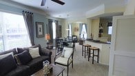 This single-floor residence showcases an open-concept layout that seamlessly connects the living, dining, and kitchen areas, creating a spacious and cohesive environment. The living area is anchored by a plush dark leather sofa, complemented by elegant accent chairs with cream upholstery, and grounded by a decorative area rug. A ceiling fan overhead and a classic table lamp add both function and warmth to the space. Large windows dressed with patterned curtains allow natural light to filter through, enhancing the soft blue and warm yellow wall tones that define the color palette throughout. The dining area sits adjacent to the living space, furnished with a dark wood table and matching chairs, and is illuminated by a pendant light fixture that adds a refined touch. A decorative wall arrangement and framed artwork contribute to the curated aesthetic. The kitchen features a breakfast bar with wicker-seat bar stools, open shelving, and dark countertops, making it both practical and visually appealing. Crown molding and arched architectural details add character and a sense of craftsmanship throughout. The neutral carpeted flooring ties all areas together, while the overall design balances comfort and style in a thoughtfully arranged living space.