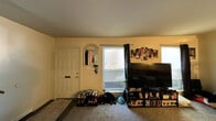This single-floor home offers a well-organized layout across several distinct living areas. The front entry opens into a carpeted living space where a front door with a mail slot is flanked by a decorative arched mirror and a key hook station, providing a practical and welcoming arrival point. Natural light filters through horizontal-blind windows, brightening the warm yellow-toned walls throughout.

The main living room is generously sized, accommodating a large sectional sofa centered around a wooden coffee table. Wall-mounted artwork and framed photo collages personalize the space, while a freestanding floor lamp adds ambient lighting. A dedicated media corner features a large flat-screen television atop an entertainment unit stocked with collectibles and media.

The kitchen is efficiently designed with dark wood-grain cabinetry, subway tile backsplash, and a full suite of stainless steel appliances including a gas range with over-range microwave and a full-size refrigerator. Dark vinyl plank flooring grounds the space with a modern aesthetic.

Adjacent to the kitchen, the dining area features hardwood-style flooring, a round natural wood dining table with matching chairs, and a large sliding glass door that floods the room with natural light and offers views of a lush green outdoor yard. A decorative ceiling fixture and ornate framed mirror complete the room's warm, inviting character.