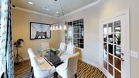 This single-floor space centers around a sophisticated dining room that blends contemporary design with eclectic touches. The room is anchored by a sleek glass-top dining table surrounded by tufted white upholstered chairs with nailhead trim detailing, creating an elegant yet inviting atmosphere. A striking zebra-print area rug adds a bold, dramatic flair underfoot and serves as a focal point for the room's aesthetic.

Overhead, a modern linear chandelier with distinctive glass pendant clusters hangs above the dining table, casting warm, ambient light throughout the space. The ceilings feature classic crown molding and a subtle tray detail, adding architectural refinement. Recessed lighting complements the chandelier for a layered lighting scheme.

One wall showcases a large mirrored grid panel that visually expands the space and connects it to an adjacent room, alongside a French door with matching grid-pane detailing. Natural light filters in through a window dressed with decorative patterned drapes in teal and brown tones over white horizontal blinds. Bold black-and-white urban photography — including iconic imagery of a Ferris wheel and the Brooklyn Bridge — adorns the walls, lending an artistic, cosmopolitan character to the overall design.