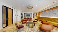 Located at 1650 Lincoln Blvd in Santa Monica, California, this single-floor space offers a refined and inviting atmosphere with a cohesive blend of modern and mid-century-inspired design. The layout includes a central lounge area, two private office rooms, and a connecting hallway, all finished with warm, natural materials and curated details.

The main lounge is an expansive gathering space featuring travertine-style tile flooring, a geometric patterned area rug, and an eclectic mix of cognac leather and velvet upholstered seating arranged around a sleek round coffee table. A sculptural pendant light anchors the ceiling, while floor-to-ceiling glass walls flood the space with natural light and offer views of a lush outdoor courtyard. A long, built-in banquette with mustard-toned upholstery lines one wall, complemented by accent tables and recessed lighting.

An adjacent media and kitchenette area features a striking oval white stone-topped island with warm wood base, leather counter stools, a wall-mounted display screen, and a tiled wet bar with dark cabinetry—ideal for presentations or casual gatherings. A bold teal abstract artwork adds a vibrant focal point.

The private offices are bright and focused, with natural light, minimalist oak desks, and tasteful wall art, creating a calm and productive environment throughout.