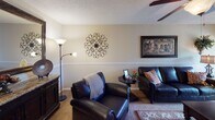 This single-floor residence offers a thoughtfully arranged layout that balances comfort and functionality across its key living areas. The living room serves as the heart of the home, featuring plush carpeted flooring, a textured ceiling, a ceiling fan, and a chair rail that runs along the walls, lending a refined architectural touch. Large vertical blinds frame a sliding door, allowing natural light to filter into the space. Adjacent to the living room, the dining area continues the warm aesthetic with rich hardwood-style flooring, a classic wooden dining table, and wall art that adds character to the space. A beam detail at the ceiling transition subtly defines the two areas. The galley-style kitchen is efficiently designed with white cabinetry, dark granite-look countertops, a double sink, white appliances including a stovetop range and refrigerator, and hardwood-style flooring, with a louvered pantry door providing additional storage. The bedroom is a serene retreat, complete with soft carpeting, a ceiling fan with light fixture, a window with decorative curtains, and ample space for dark wood furniture. The full bathroom features a tub-shower combination with tile surround, dark countertop vanity, and hardwood-style flooring, completing this cohesive and welcoming home.