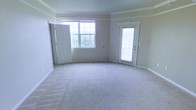 This single-floor space offers a clean, well-maintained interior designed with comfort and functionality in mind. The main living area is generously proportioned, featuring soft, neutral-toned wall-to-wall carpeting that adds warmth underfoot. Crown molding lines the ceiling throughout, lending a classic architectural detail that elevates the overall finish of the space.

Natural light is a defining characteristic of this space. A large multi-pane window allows abundant daylight to pour in, brightening the interior and creating an airy, open atmosphere. A glass-panel exterior door further enhances the light-filled quality of the room while providing direct outdoor access. The neutral wall tones complement the light carpeting, creating a cohesive and understated palette that would suit a variety of interior styles.

The layout is open and unobstructed, offering flexible possibilities for furniture arrangement and use of the space. Doorways are framed with clean white trim, consistent with the traditional detailing found throughout. The combination of crown molding, multi-pane windows, and classic door surrounds gives the space a timeless, traditional character. Overall, this is a bright, move-in-ready space with thoughtful architectural details and a welcoming ambiance.