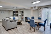 Located at 137 N Orange Ave in Brea, California, this single-floor space offers a thoughtfully designed open-plan layout that seamlessly integrates multiple functional areas into one cohesive environment. The interior features warm-toned luxury vinyl plank flooring throughout, providing a clean and consistent foundation that enhances the overall brightness of the space.

The dining area is anchored by a large round wood-finished table surrounded by upholstered navy blue chairs, positioned adjacent to a floor-to-ceiling vertical blind window treatment that allows for generous natural light and privacy control. Floral-patterned curtains add a soft decorative touch to the windows.

The living and lounge area features a substantial gray sectional sofa paired with a round wooden coffee table, all oriented near a classic fireplace that serves as a focal point along the back wall. Two built-in dark-finished bookshelves flank a recessed nook and are filled with books, adding a warm, intellectual character to the space.

A separate seating nook accommodates two houndstooth-patterned wingback chairs accented with teal and yellow throw pillows, creating an inviting reading or conversation corner. Overhead, large oval flush-mount light fixtures provide even, ambient illumination throughout. The space is well-ventilated with ceiling HVAC vents and presents a clean, comfortable atmosphere suited for community gathering or shared living.