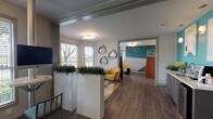 This single-floor media room is a thoughtfully designed communal space that balances collaborative function with contemporary style. The layout is organized into several distinct zones, each serving a specific purpose while maintaining a cohesive aesthetic throughout. Rich wood-look flooring runs continuously across the space, anchoring the various areas with warmth and continuity.

A comfortable lounge area anchors one end of the room, featuring a bay window configuration that allows generous natural light and outdoor views. Four mustard-yellow accent chairs are arranged around low round side tables atop a chevron-patterned area rug, creating an inviting setting for casual conversation or relaxation.

Adjacent to the lounge, a glass-partitioned workspace provides a semi-private area with bar-height tables and seating, accented by an olive-yellow feature wall and curated artwork. The transparency of the glass enclosure maintains visual openness while offering a degree of separation.

A dedicated conference area showcases a bold geometric mural in tones of gold, teal, lavender, and white. A round white table surrounded by upholstered rolling chairs sits beneath a dramatic multi-bulb chandelier, making it well-suited for group meetings or presentations.

Decorative geode-style wall art, tall white planters with greenery, and a beverage station with a wine cooler complete the space, adding personality and everyday convenience throughout.
