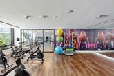 Located at 1015 Spring Garden Rd in Miami, FL, this well-appointed fitness studio offers a thoughtfully designed space for a comprehensive workout experience. The studio features warm hardwood-style flooring throughout, clean white ceilings with recessed linear LED lighting, and a bright, modern aesthetic that creates an energizing atmosphere.

The main workout area is anchored by a row of professional spin bikes positioned directly in front of expansive floor-to-ceiling windows, providing inspiring views of a lush, tropical poolside courtyard with palm trees and resort-style lounge seating. This outdoor connection floods the interior with natural light and adds a distinctly Miami ambiance to every session.

A large-format motivational mural depicting athletes in action serves as a dynamic focal point on one wall, complementing an organized rack stocked with stability balls and color-coded medicine balls. Full-length mirrors along the walls enhance the sense of space and support proper form during training.

Glass-paneled doors connect this studio to an adjacent strength and cardio equipment room, allowing for seamless transitions between workout zones. Additional amenities include a built-in credenza, a wall-mounted television, and vibrant tropical artwork that reinforces the space's energetic, Miami-inspired character. The studio is well-ventilated with ceiling-integrated HVAC and is designed to support group fitness classes as well as individual training.