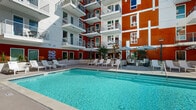 Located on Santa Monica Boulevard in the heart of West Los Angeles, this outdoor courtyard amenity space offers a resort-inspired experience defined by striking contemporary architecture. The building's bold facade — a vivid combination of deep terracotta orange and crisp white paneling — frames the courtyard on multiple sides, rising several stories with private balconies that overlook the pool area below.

At the center of the space is a generously sized swimming pool with clear, turquoise water and clean tile detailing, complemented by a separate spa. The expansive pool deck is outfitted with white lounge chairs and shaded by navy-and-white striped umbrellas, providing ample space for relaxation. A dedicated cabana area features elegant wood-framed daybeds draped with flowing white curtains, adding a luxurious, retreat-like atmosphere.

Raised planting beds with lush greenery and tropical plantings line portions of the deck, softening the architectural hardscape and adding a natural element to the environment. Comfortable seating vignettes, including hanging egg chairs, are thoughtfully arranged throughout the courtyard, creating inviting gathering spots. The open-air layout captures abundant Southern California sunlight, making this a bright, vibrant outdoor destination ideal for leisure and socializing.