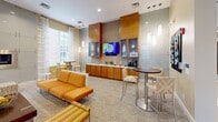 Located at 1982 State Highway 70 E in Cherry Hill, NJ, this thoughtfully designed single-floor communal space offers a welcoming and stylish environment for gathering and relaxation. The layout is open and versatile, accommodating multiple seating configurations across distinct zones that flow seamlessly into one another.

At the heart of the space sits a sophisticated lounge area anchored by a sleek, wall-mounted linear fireplace set against a frosted glass panel backdrop — a striking architectural focal point. Bold orange sofas and accent chairs arranged symmetrically around a dark wood coffee table create a warm, inviting atmosphere. Patterned carpet in neutral tones adds texture underfoot throughout the main areas.

Natural light pours in generously through multiple large windows dressed with sheer, striped curtains, lending the space an airy, open feel with views of the surrounding greenery. A cozy conversation nook with upholstered chairs and side tables provides a quieter retreat within the broader space.

A dedicated café-style area features warm wood cabinetry, a mounted flat-screen display, pendant lighting, and counter-height seating — ideal for casual meetings or a morning coffee. The entry foyer transitions elegantly from polished tile flooring into the carpeted interior, with glass-paneled walls, a modern chandelier, and framed artwork adding refined finishing touches throughout.