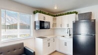 This efficiently designed 2-bedroom, 1-bathroom single-story unit offers a thoughtfully arranged open-concept layout that maximizes every square foot. Light-toned luxury vinyl plank flooring runs throughout, complemented by crisp white walls and recessed lighting that keep the interior feeling bright and airy.

The kitchen is a standout feature, showcasing white shaker-style cabinetry with matte black hardware, a subway tile backsplash, a built-in microwave, a smooth cooktop, a deep single-basin sink, and a full-size refrigerator. Decorative greenery atop the upper cabinets adds a lively touch. The kitchen flows seamlessly into the living area, creating an inviting open space ideal for everyday living.

Both bedrooms are clean and versatile, featuring white walls, recessed lighting, and natural light from windows with horizontal blinds. The primary bedroom includes a built-in wardrobe cabinet with matching matte black hardware, offering practical storage. The second bedroom provides a blank canvas with ample open floor space.

The bathroom is a refined retreat, finished with marble-look wall tile, a glass-enclosed walk-in shower with a built-in bench and recessed niches, a dual-sink white vanity with a quartz countertop, matte black fixtures, and a large mirror — all lending a polished, contemporary aesthetic throughout.