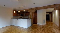 This single-floor space masterfully combines historic character with contemporary design. Exposed, heavy timber beams serve as a defining architectural feature throughout, adding warmth and a sense of heritage to the interior. Light hardwood floors run continuously across the space, enhancing the bright, airy atmosphere created by white walls and natural light filtering in through windows at the far end of the hallway.

The corridor leading through the space is framed by substantial wooden post-and-beam construction, lending an industrial-loft aesthetic. Track lighting mounted along the ceiling provides adjustable illumination throughout.

The kitchen is well-appointed, featuring rich dark wood cabinetry that contrasts beautifully with the lighter surroundings. A peninsula with a granite countertop and bar seating serves as both a functional workspace and a social hub. Stainless steel appliances, including a refrigerator and microwave, add a modern touch, while pendant lights hang above the peninsula, creating a warm, inviting ambiance. A prominent timber post anchors the kitchen area, reinforcing the space's distinctive rustic-meets-modern character.