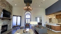 This thoughtfully designed single-floor space makes a striking first impression with its soaring vaulted ceilings and dramatic architectural details. At the heart of the room hangs a statement antler chandelier, adding a rustic yet refined focal point that draws the eye upward and highlights the impressive ceiling height. A grand arched entryway frames a set of glass double doors, flooding the interior with abundant natural light and providing an open, welcoming feel. The tray ceiling detail, finished in a deep contrasting tone, adds architectural depth and visual interest overhead. The main lounge area is furnished with contemporary grey sofas and armchairs, complemented by a sleek glass coffee table and a modern floor lamp, creating a comfortable and stylish gathering space. Dark hardwood flooring anchors the room and contrasts beautifully with the crisp white walls. An adjacent room, visible through a large mirrored opening, appears to offer additional seating and dining, extending the functionality of the overall space. The combination of rustic elements, such as the antler chandelier, with clean modern furnishings and abundant natural light gives this space a warm, inviting ambiance well-suited for socializing and relaxation.