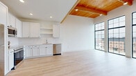 This thoughtfully designed 2-bedroom, 2-bathroom residence in Ankeny, Iowa, delivers a striking blend of contemporary style and functional living. The open-concept kitchen and living area is an immediate standout, featuring soaring ceilings with warm wood-paneled detailing, recessed lighting, and light-toned hardwood flooring that flows seamlessly throughout. The kitchen is well-appointed with white shaker cabinetry, stainless steel appliances including a refrigerator, range, microwave, and dishwasher, plus open shelving and clean white countertops. Dramatic floor-to-ceiling black-framed windows and French doors flood the living space with abundant natural light and open to two private balconies, offering expansive views of the surrounding Ankeny streetscape. The bedrooms are bright and airy, each featuring closets and clean, minimalist finishes. Both bathrooms are finished with white vanities, LED-backlit mirrors, black hardware accents, and light hardwood-style flooring, maintaining a cohesive modern aesthetic. Additional conveniences include an in-unit laundry and a hallway connecting the living areas. The overall ambiance is clean, contemporary, and light-filled, making excellent use of its well-proportioned layout.