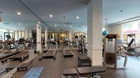 Located at 65 N Madison Ave in Pasadena, California, this fitness center offers a comprehensive, single-level workout facility designed to accommodate a wide range of exercise needs. The space is thoughtfully divided into distinct training zones, each outfitted with purpose-driven equipment.

The cardio area is anchored by a row of Matrix treadmills and elliptical machines, positioned beneath recessed lighting and flanked by floor-to-ceiling mirrors that visually expand the space. Rubber flooring throughout provides durability and safety underfoot.

A dedicated strength and functional training zone features a variety of resistance machines, including leg press and cable systems, alongside free weights, dumbbells, stability balls, and exercise mats labeled for power, balance, and endurance training. This area benefits from natural light streaming through large windows that offer views of the surrounding outdoor greenery and courtyard.

A separate glass-walled room houses additional strength machines and a cable weight system, providing a more focused environment for targeted resistance training. Structural columns punctuate the layout while maintaining an open, airy feel throughout.

The facility's neutral color palette, combination of recessed and pendant lighting, and mirrored surfaces contribute to a clean, professional ambiance well-suited for daily fitness routines.