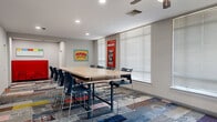 This energetic, single-floor collaborative workspace is thoughtfully designed to inspire productivity and creativity. The space is unified throughout by a striking patchwork carpet in vivid blocks of blue, red, pink, and grey, setting a bold and dynamic tone from the moment you enter.

The main conference area serves as the heart of the space, centered around a large communal high-top table surrounded by sleek navy chairs — ideal for group meetings, brainstorming sessions, or collaborative work. Natural light filters in through multiple windows fitted with horizontal blinds, brightening the grey-walled interior. Eye-catching pop-art-inspired wall pieces, including a bold "WKORN" comic panel and a red telephone booth-style motivational print reading "Talk Less, Do More," add personality and visual energy.

A smaller, private study room offers a more intimate setting, equipped with a square table, vibrant lime-green chairs, a large wall-mounted whiteboard, and an encouraging "The Answer Is Simple" chalkboard-style artwork — perfect for focused group study or small meetings.

A comfortable lounge area rounds out the space, featuring a bold blue sofa flanked by side tables and adorned with motivational typographic art pieces. Ceiling fans are present throughout, and a glazed interior door connects the lounge to an adjacent area, enhancing the open, connected feel of this well-appointed collaborative environment.