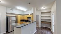 This single-story residence offers a well-designed, functional layout that balances open communal spaces with private living areas. The kitchen serves as a central highlight, featuring warm honey-toned cabinetry, dark gray countertops, and a full suite of stainless steel appliances including a refrigerator, electric range, microwave, and dishwasher. A peninsula with an extended countertop provides additional prep space and separates the kitchen from the rest of the living area. Pendant lighting and a built-in shelving nook add both style and practicality to this space. Rich, dark hardwood-style flooring extends throughout the kitchen and main living areas, lending warmth and cohesion to the interior. The bedrooms are carpeted for comfort and each features a window that allows natural light to filter in. One bedroom includes a ceiling fan with light fixture. The primary bathroom is equipped with a full tub-shower combination framed by white tile, while a second bathroom offers a vanity with cabinetry and matching countertop. The neutral gray and white color palette throughout creates a clean, modern aesthetic that is both inviting and versatile.