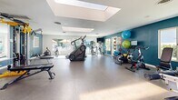 Located at 2401 S Hacienda Blvd in Hacienda Heights, CA, this single-story fitness center offers a thoughtfully designed space for comprehensive workouts. The main gym area is bright and open, bathed in natural light streaming through large skylights and windows that provide views of the outdoor surroundings. The durable speckled flooring throughout the workout space is well-suited for high-traffic athletic use. The gym is fully equipped with a wide range of cardio machines, including treadmills, ellipticals, a stair climber, and stationary cycling bikes. A dedicated strength training section features a cable machine, free weights with a rack, and a weight bench, complemented by wall-mounted mirrors that enhance the sense of space. Exercise balls and additional accessories are neatly organized on wall-mounted storage. Teal accent walls add a vibrant, energizing aesthetic throughout the space, creating a motivating atmosphere. The accessible bathroom is clean and functional, featuring warm wood-look vinyl flooring, teal accent walls consistent with the gym's design, ADA-compliant grab bars, a wall-mounted sink, and a well-lit vanity mirror. A connecting hallway links the facility's rooms together, ensuring smooth and accessible flow throughout the space.