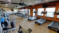This fitness center offers a thoughtfully designed workout environment built to accommodate a wide variety of exercise needs. The space is finished with warm terracotta-toned accent walls that create an energizing atmosphere, complemented by durable, textured carpet flooring suited for high-traffic use. Full-length mirrors line one wall, enhancing the sense of openness while allowing users to monitor their form during exercise. Natural light flows in through multiple windows fitted with blinds, keeping the space bright and welcoming. Overhead ceiling fans provide additional air circulation throughout the room. The cardio section features a lineup of treadmills, providing ample space for multiple users simultaneously. A well-stocked free weights area includes a rack of medicine balls and a full set of dumbbells arranged for easy access. Strength training equipment is also present, rounding out the facility's comprehensive offerings. Recessed lighting and mounted televisions add to the functionality and comfort of the space. An emergency exit door ensures safety compliance, while the overall layout promotes ease of movement between equipment zones. This fitness center is designed to deliver a complete and convenient exercise experience.