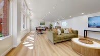Located at 1942 Deerpark Dr in Fullerton, California, this thoughtfully designed single-floor community living room offers a welcoming and versatile gathering space. The open layout is anchored by light-toned hardwood flooring that runs throughout, complementing the crisp white walls and recessed lighting that keeps the space bright and airy.

Large floor-to-ceiling windows along one wall flood the room with natural light and offer sweeping views of an outdoor pool area and pergola, creating a seamless connection between interior and exterior. A seating area features a plush olive-green sofa accented with patterned throw pillows and arranged around a textured drum coffee table, providing a relaxed lounge atmosphere.

Multiple round marble-top dining tables with sleek black bases and cognac leather chairs are distributed throughout the space, accommodating group gatherings with ease. A wall-mounted flat-screen TV paired with a contemporary console table and lush potted fiddle-leaf fig trees add functional and decorative elements throughout. A mid-century-style credenza and curated wall art, including a vibrant landscape painting, contribute warmth and character to the overall design. A wall-mounted mini-split system ensures year-round comfort. The result is a bright, modern, and inviting communal space ideal for socializing and relaxation.