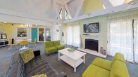 Located at 8470 Towson Blvd. in Miamisburg, Ohio, this single-floor community clubhouse offers a welcoming and vibrantly designed gathering space. The main lounge area is anchored by dramatic vaulted ceilings with exposed white structural trusses and skylights that flood the room with natural light. A brick fireplace with a wall-mounted flat-screen TV serves as the focal point of the lounge, which is furnished with contemporary chartreuse sofas, a tufted leather sectional, and a clean white coffee table — all set atop a patterned carpet floor. Sheer white curtains frame large windows, softening the natural light and adding an airy feel to the space. Adjacent to the lounge is a flexible meeting and gathering area featuring a round white table with modern yellow chairs, a storage credenza, and colorful artwork on the walls. A bold teal accent color carries throughout the space, appearing on doors, walls, and architectural details, giving the clubhouse a cohesive, energetic aesthetic. The kitchenette and bar area features a curved dark countertop with bar seating, pendant lighting, a pass-through window, and full kitchen appliances — ideal for hosting events and social gatherings. Overall, the space presents a lively, modern atmosphere suited for community engagement and leisure.
