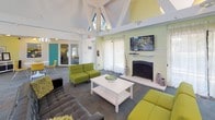 Located at 8470 Towson Blvd. in Miamisburg, Ohio, this single-floor community clubhouse offers a welcoming and vibrantly designed gathering space. The main lounge area is anchored by dramatic vaulted ceilings with exposed white structural trusses and skylights that flood the room with natural light. A brick fireplace with a wall-mounted flat-screen TV serves as the focal point of the lounge, which is furnished with contemporary chartreuse sofas, a tufted leather sectional, and a clean white coffee table — all set atop a patterned carpet floor. Sheer white curtains frame large windows, softening the natural light and adding an airy feel to the space. Adjacent to the lounge is a flexible meeting and gathering area featuring a round white table with modern yellow chairs, a storage credenza, and colorful artwork on the walls. A bold teal accent color carries throughout the space, appearing on doors, walls, and architectural details, giving the clubhouse a cohesive, energetic aesthetic. The kitchenette and bar area features a curved dark countertop with bar seating, pendant lighting, a pass-through window, and full kitchen appliances — ideal for hosting events and social gatherings. Overall, the space presents a lively, modern atmosphere suited for community engagement and leisure.