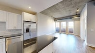 This one-bedroom, one-bathroom urban loft showcases an industrial-modern aesthetic defined by striking exposed concrete ceilings with visible ductwork and track lighting throughout. The open-concept layout seamlessly connects the kitchen and living area, creating a spacious, airy feel. The kitchen is well-appointed with white shaker cabinetry, dark granite countertops, a subway tile backsplash, and a full suite of stainless steel appliances including a French door refrigerator, electric range, and over-the-range microwave. A large center island provides additional prep and dining space. The living area is bathed in natural light from floor-to-ceiling glass doors that open directly onto a private balcony, offering views of the surrounding cityscape. Light-toned hardwood flooring runs throughout the main living spaces, while the bedroom features the same warm wood floors and a distinctive exposed concrete column, adding to the loft's character. A walk-in closet with built-in shelving and hanging rods provides ample storage. The bathroom is finished with a white vanity, dark granite countertop, tile flooring, and a glass-enclosed shower. A stacked in-unit washer and dryer adds everyday convenience to this thoughtfully designed urban residence.