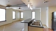 This single-floor space showcases a thoughtfully designed open-concept layout that seamlessly connects the kitchen and living areas. The kitchen is anchored by a sleek island with a dark countertop and a double stainless steel sink, complemented by warm wood cabinetry underneath. A striking cluster of cylindrical pendant lights hangs above the island, adding a modern focal point to the space. The living area is finished with carpeted flooring and benefits from ample natural light streaming through multiple windows fitted with horizontal blinds. A ceiling fan serves the living area, providing both comfort and a casual aesthetic touch. Adding an industrial character to the space, an exposed metallic duct runs prominently along the ceiling, creating an urban, loft-inspired feel. The neutral wall tones and clean lines throughout contribute to a bright, airy ambiance. The open flow between the kitchen and living space makes the overall layout feel expansive and functional, well-suited for everyday living and entertaining alike.