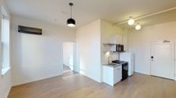 This single-floor space offers a bright and modern open-concept layout designed for efficient, comfortable living. The interior is finished with light hardwood flooring that runs throughout, creating a warm and cohesive aesthetic. Crisp white walls and high ceilings amplify the sense of openness and allow natural and artificial light to reflect evenly across the space.

The compact kitchen is thoughtfully arranged and tucked along one wall, featuring white cabinetry, open shelving, a stainless steel sink, a gas range, and a microwave. A full-size refrigerator rounds out the kitchen's functionality, making the most of the available footprint. Pendant lighting above the main living area adds a contemporary touch, with a sleek black dome fixture serving as a design focal point.

A partial dividing wall separates the kitchen from the main living area, providing a sense of structure without sacrificing the open feel of the floor plan. A doorway leads to an additional room beyond, offering a degree of separation and privacy. The overall ambiance is clean, minimal, and move-in ready, with a neutral palette that provides a versatile backdrop for a variety of interior styles.