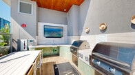 Located at 110 Beech Street in the heart of San Diego, this rooftop amenity deck offers a sophisticated outdoor retreat with panoramic views of the city skyline. The space is defined by clean, modern architecture featuring smooth concrete surfaces, warm wood accents, and carefully curated landscaping throughout.

At the center of the deck is a fully equipped outdoor kitchen and lounge area, sheltered beneath a striking wood-paneled ceiling. The kitchen features dual stainless steel grills, a utility sink, and a wall-mounted flat-screen television, all finished with distinctive green tilework that adds a vibrant design touch. Grill instructions are posted for resident convenience, making this an ideal space for entertaining.

Stepping out onto the open terrace, expansive concrete planters overflow with lush greenery, tropical plants, and shrubs, bringing a garden-like atmosphere to the rooftop. Glass railings and metal balustrades frame the perimeter, ensuring unobstructed views of the San Diego skyline and surrounding neighborhoods under the abundant Southern California sun.

Comfortable lounge seating, including wicker chairs and side tables, is arranged to take full advantage of the open-air environment. Decorative wood slat screening adds architectural texture to the exterior walls, while ambient wall-mounted lighting enhances the space for evening use. The overall ambiance is modern, airy, and thoughtfully designed for relaxation and social gatherings.