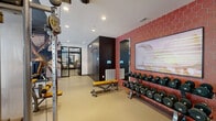 This modern fitness center is thoughtfully designed to support a full range of workout routines across a generous, open floor plan. The space is divided into distinct functional zones, each tailored to a specific type of training. The cardio area lines the floor-to-ceiling windows, where TRUE and Life Fitness treadmills, ellipticals, and stationary bikes are positioned to take advantage of abundant natural light and exterior views of the surrounding neighborhood. A dedicated strength training section features multi-station cable machines, a dumbbell rack with a full range of weights, and a flat bench, all set against durable rubberized flooring. The studio zone transitions to hardwood-style flooring and includes ICG indoor cycling bikes, TRX suspension trainers, stability balls, and a ballet barre, with large framed mirrors and a glass door entry enhancing the open feel. Design elements include an exposed industrial ceiling with track and recessed lighting, bold hexagonal red tile accent walls, and curated artwork. A refrigerated beverage station is also visible, adding a practical amenity. The overall ambiance is modern, energetic, and professional, combining industrial aesthetics with high-end fitness amenities.