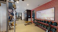 This modern fitness center is thoughtfully designed to support a full range of workout routines across a generous, open floor plan. The space is divided into distinct functional zones, each tailored to a specific type of training. The cardio area lines the floor-to-ceiling windows, where TRUE and Life Fitness treadmills, ellipticals, and stationary bikes are positioned to take advantage of abundant natural light and exterior views of the surrounding neighborhood. A dedicated strength training section features multi-station cable machines, a dumbbell rack with a full range of weights, and a flat bench, all set against durable rubberized flooring. The studio zone transitions to hardwood-style flooring and includes ICG indoor cycling bikes, TRX suspension trainers, stability balls, and a ballet barre, with large framed mirrors and a glass door entry enhancing the open feel. Design elements include an exposed industrial ceiling with track and recessed lighting, bold hexagonal red tile accent walls, and curated artwork. A refrigerated beverage station is also visible, adding a practical amenity. The overall ambiance is modern, energetic, and professional, combining industrial aesthetics with high-end fitness amenities.