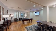 The resident lounge is a well-appointed communal space designed for both relaxation and recreation. The room's centerpiece is a classic billiards table positioned atop a stylish area rug, offering residents a go-to spot for entertainment. Warm hardwood flooring extends throughout the space, adding a welcoming, natural aesthetic that complements the neutral-toned walls and recessed lighting overhead.

A wall-mounted flat-screen television anchors one side of the room, ideal for gathering to watch sports or movies. Nearby, a dining-height table paired with industrial-style metal chairs provides a casual setting for games or casual meals. On the opposite side, a comfortable seating arrangement with upholstered chairs and floor lamps creates a quieter lounge area perfect for conversation or unwinding.

Large windows along one wall allow natural light to filter into the space, enhancing the open, airy feel. The combination of recreational features, flexible seating zones, and thoughtful design elements makes this lounge a versatile and inviting amenity for everyday use.