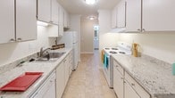 This upgraded one-bedroom, one-bathroom residence is thoughtfully designed across a single floor, offering a comfortable and functional layout. The galley-style kitchen is well-appointed with white cabinetry, granite-style countertops, under-cabinet lighting, and white appliances including a four-burner electric range, refrigerator, and dishwasher. Tile flooring runs throughout the kitchen, which connects seamlessly to the rest of the unit. The living area is generously sized, featuring soft carpet flooring, crown molding, and neutral warm-toned walls that create a welcoming ambiance. A sliding glass door opens to a private balcony, framing views of lush greenery and providing an abundance of natural light. The bedroom is a peaceful retreat, also carpeted and accented with crown molding, and benefits from a window overlooking verdant outdoor scenery. The full bathroom is clean and modern, showcasing a white vanity with an integrated sink, a mirrored medicine cabinet, tile flooring, and a bathtub with a glass sliding door enclosure. Throughout the unit, consistent crown molding and neutral tones contribute to a cohesive, polished aesthetic.