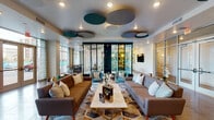 This thoughtfully designed single-floor communal amenity space combines modern aesthetics with practical functionality across several well-appointed areas. The main lounge is anchored by a pair of warm brown mid-century sofas flanking a marble-topped coffee table, set atop a vibrant geometric area rug. Overhead, a striking ceiling installation of circular acoustic panels in teal, blue, and taupe adds artistic flair while managing acoustics. A glass-partitioned enclave in the background provides a semi-private seating nook, adding depth and versatility to the lounge.

A secondary seating area features sleek gray sofas and gold-accented console tables, with a wall-mounted flat-screen TV and additional circular ceiling panels continuing the cohesive design language. Large floor-to-ceiling windows and glass entry doors flood the entrance with natural light, offering views of the surrounding streetscape and creating an open, welcoming atmosphere.

The kitchen is a standout feature, showcasing a generously sized island with a crisp white quartz countertop and tiled base, flanked by upholstered gray barstools. Dramatic gold orb pendant lights hang overhead, complementing warm wood cabinetry and a vivid teal tile backsplash. Stainless steel appliances, a wall oven, wine cooler, and a mounted TV complete this highly functional and visually impressive space.