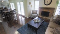 This single-story living space combines classic and casual design elements to create a warm, inviting atmosphere. The living area is anchored by rich, dark hardwood floors that extend throughout, complemented by a plush blue-gray area rug that defines the seating zone. A dark wood coffee table serves as the centerpiece, styled with decorative accents on a tray.

Natural light floods the space through multiple windows fitted with white plantation shutters, as well as a set of French doors that offer a bright, airy connection to the outdoors. The walls are finished in a soft neutral tone, providing a clean backdrop for the room's furnishings and decor, including decorative wall medallions that add a subtle artistic touch.

A brick fireplace with a raised hearth anchors one wall, serving as a striking focal point that adds character and warmth to the room. Adjacent to the living area, a glimpse of a bar-height dining setup with tall chairs is visible, suggesting a seamless transition between living and dining spaces. The overall ambiance is comfortable and well-lit, with a cohesive blend of traditional and transitional design elements.