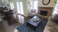 This single-story living space combines classic and casual design elements to create a warm, inviting atmosphere. The living area is anchored by rich, dark hardwood floors that extend throughout, complemented by a plush blue-gray area rug that defines the seating zone. A dark wood coffee table serves as the centerpiece, styled with decorative accents on a tray.

Natural light floods the space through multiple windows fitted with white plantation shutters, as well as a set of French doors that offer a bright, airy connection to the outdoors. The walls are finished in a soft neutral tone, providing a clean backdrop for the room's furnishings and decor, including decorative wall medallions that add a subtle artistic touch.

A brick fireplace with a raised hearth anchors one wall, serving as a striking focal point that adds character and warmth to the room. Adjacent to the living area, a glimpse of a bar-height dining setup with tall chairs is visible, suggesting a seamless transition between living and dining spaces. The overall ambiance is comfortable and well-lit, with a cohesive blend of traditional and transitional design elements.