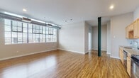 This single-floor loft-style unit blends industrial character with modern residential comfort. The open living area is immediately striking, showcasing warm hardwood flooring that extends throughout the main spaces, exposed silver ductwork running along the ceiling, and a large multi-pane industrial window that floods the room with abundant natural light and urban views. A distinctive dark green structural column adds an architectural focal point to the open layout.

The kitchen is well-appointed with light maple cabinetry, granite-style countertops, a mosaic tile backsplash, and a full suite of black appliances including a range, over-the-range microwave, dishwasher, and refrigerator — offering both functionality and style.

The bedrooms continue the industrial aesthetic, featuring plush neutral carpeting, exposed ductwork, and wide industrial-style windows with horizontal blinds that frame outdoor views. Each bedroom offers a comfortable and quiet retreat with ample wall space and good natural light.

The bathroom is clean and functional, featuring warm wood-toned cabinetry, a white countertop vanity with a large mirror, hardwood flooring, and a full soaking tub and shower combination with crisp white surrounds. The overall ambiance of the unit is open, airy, and urban.
