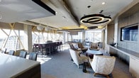 This sophisticated amenity lounge is designed to offer a dynamic and versatile social environment. The space is organized into several distinct zones, each serving a unique purpose while maintaining a cohesive, upscale aesthetic throughout.

At the heart of the lounge, a generously sized seating area features plush, neutral-toned armchairs and sofas arranged around low accent tables, creating an inviting atmosphere for relaxed conversation or casual gatherings. Striking circular pendant light fixtures hang overhead, adding a bold, modern focal point to the ceiling.

Adjacent to the lounge seating, a high-top dining area with dark wood bar-height tables and upholstered chairs provides an ideal setting for group dining or socializing. A billiards table positioned near the windows adds a recreational dimension to the space.

Floor-to-ceiling windows along the perimeter flood the interior with abundant natural light and offer expansive exterior views, enhancing the open, airy feel of the room. Geometric wood-framed architectural detailing frames the windows, lending a distinctive and contemporary design element.

The flooring features neutral carpeting that complements the warm, muted color palette of the furnishings and finishes. Recessed lighting supplements the pendant fixtures, ensuring the space is well-lit at all times. Overall, the lounge strikes a refined balance between comfort and modern design.