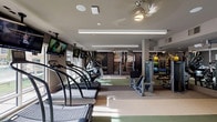 This fitness center in Houston, TX offers a comprehensive, single-floor workout facility designed to accommodate a wide range of training needs. The space is organized into several distinct zones, each tailored to specific exercise disciplines. The cardio area features a row of treadmills—including Woodway models—alongside ellipticals and a stair climber, all positioned near large windows that allow natural light to filter in and provide views of the outdoors. Wall-mounted televisions are positioned throughout for entertainment during workouts.

The strength training section is well-appointed with cable machines, selectorized equipment, and a dedicated free weights area featuring a full dumbbell rack and adjustable benches, complemented by large mirrors along the walls. A separate cycling alcove houses multiple stationary bikes on a cushioned floor surface, benefiting from additional natural light through surrounding windows.

The flooring throughout transitions between rubberized and carpeted surfaces depending on the zone, providing both durability and comfort. Overhead illumination is provided by a combination of recessed lighting and linear LED fixtures, while ceiling fans contribute to air circulation. Motivational athletic murals and bold graphic wall art add visual energy to the environment. The overall design is modern and functional, creating a well-organized and inviting space for fitness training.