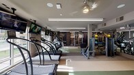 This fitness center in Houston, TX offers a comprehensive, single-floor workout facility designed to accommodate a wide range of training needs. The space is organized into several distinct zones, each tailored to specific exercise disciplines. The cardio area features a row of treadmills—including Woodway models—alongside ellipticals and a stair climber, all positioned near large windows that allow natural light to filter in and provide views of the outdoors. Wall-mounted televisions are positioned throughout for entertainment during workouts.

The strength training section is well-appointed with cable machines, selectorized equipment, and a dedicated free weights area featuring a full dumbbell rack and adjustable benches, complemented by large mirrors along the walls. A separate cycling alcove houses multiple stationary bikes on a cushioned floor surface, benefiting from additional natural light through surrounding windows.

The flooring throughout transitions between rubberized and carpeted surfaces depending on the zone, providing both durability and comfort. Overhead illumination is provided by a combination of recessed lighting and linear LED fixtures, while ceiling fans contribute to air circulation. Motivational athletic murals and bold graphic wall art add visual energy to the environment. The overall design is modern and functional, creating a well-organized and inviting space for fitness training.