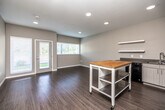 This single-floor unit presents a clean, contemporary design with a neutral color palette of warm grays and soft whites throughout. The open-concept living and kitchen area is anchored by durable wood-look plank flooring and benefits from generous natural light through multiple windows fitted with white horizontal blinds. A glass-panel exterior door connects the living space to the outdoors, enhancing the bright, airy atmosphere.

The kitchen is well-equipped with light-toned cabinetry, black appliances including a range, over-the-range microwave, refrigerator, and dishwasher, along with a textured countertop and single-basin sink. Open wall shelving adds additional storage, while a mobile butcher block island provides flexible workspace and extra prep area.

The bedroom features plush carpeting, a ceiling fan, a wide window with outdoor views, and a full-width closet with wire shelving for organized storage.

The bathroom is finished with white hexagonal tile flooring, a sleek white vanity with an extended countertop, a large mirror, and a stacked washer and dryer unit conveniently integrated into the same space. A textured ceiling and recessed lighting appear throughout, contributing to the unit's modern, functional character.